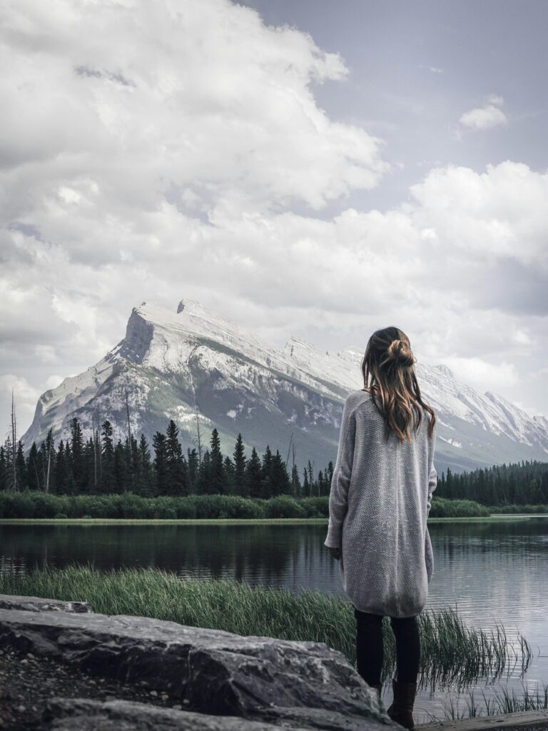 A contemplative woman stands by a tranquil lake with a mountain backdrop under a cloudy sky.