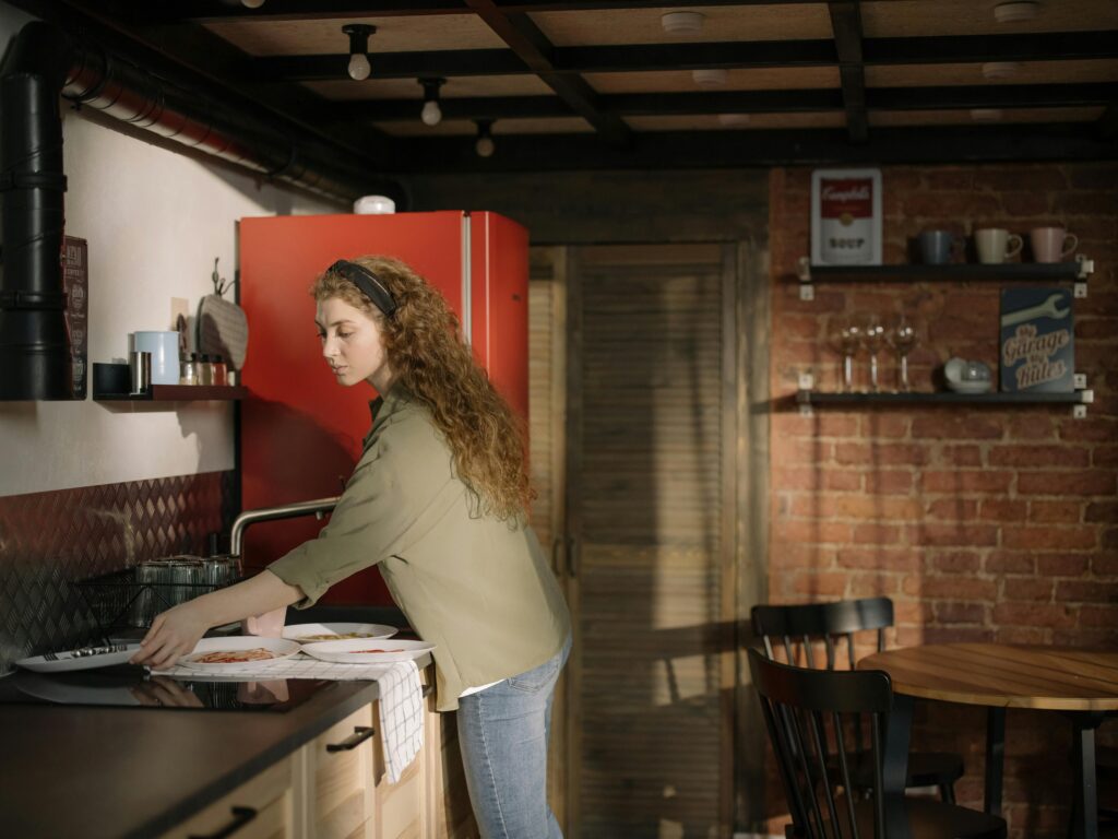 A woman arranging dinnerware in a cozy kitchen with a red fridge and brick wall.