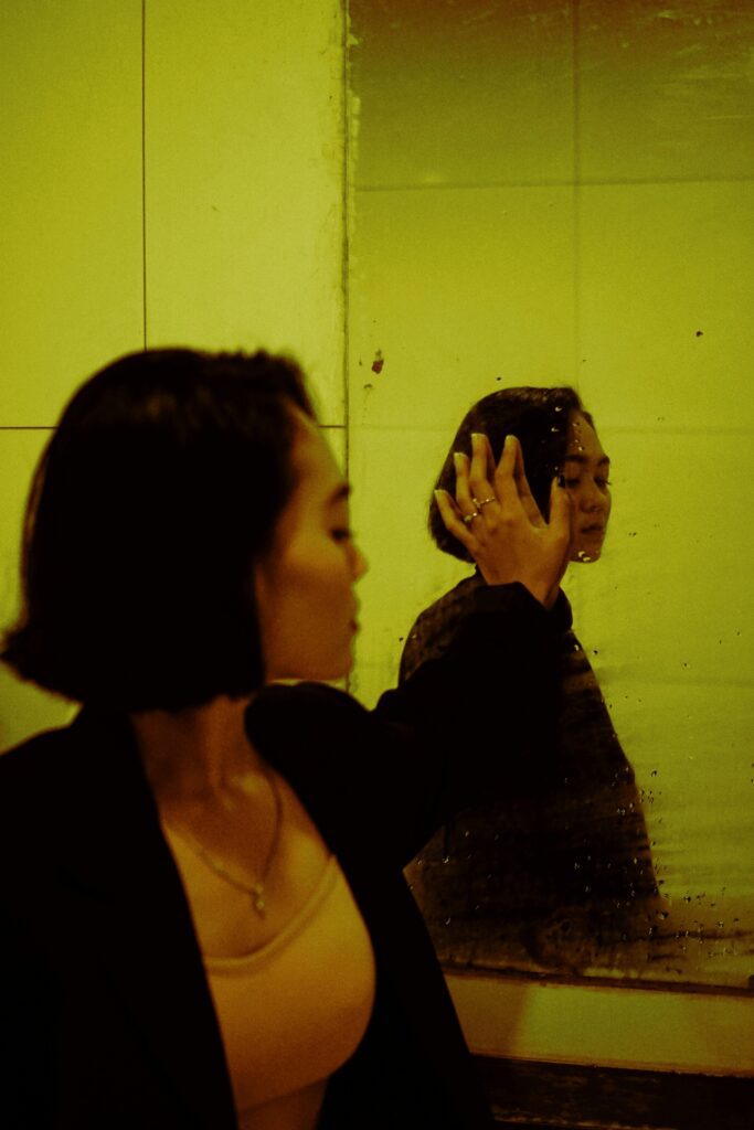 Side view of young depressed ethnic female in jacket reflecting in mirror with water drops while looking away indoors