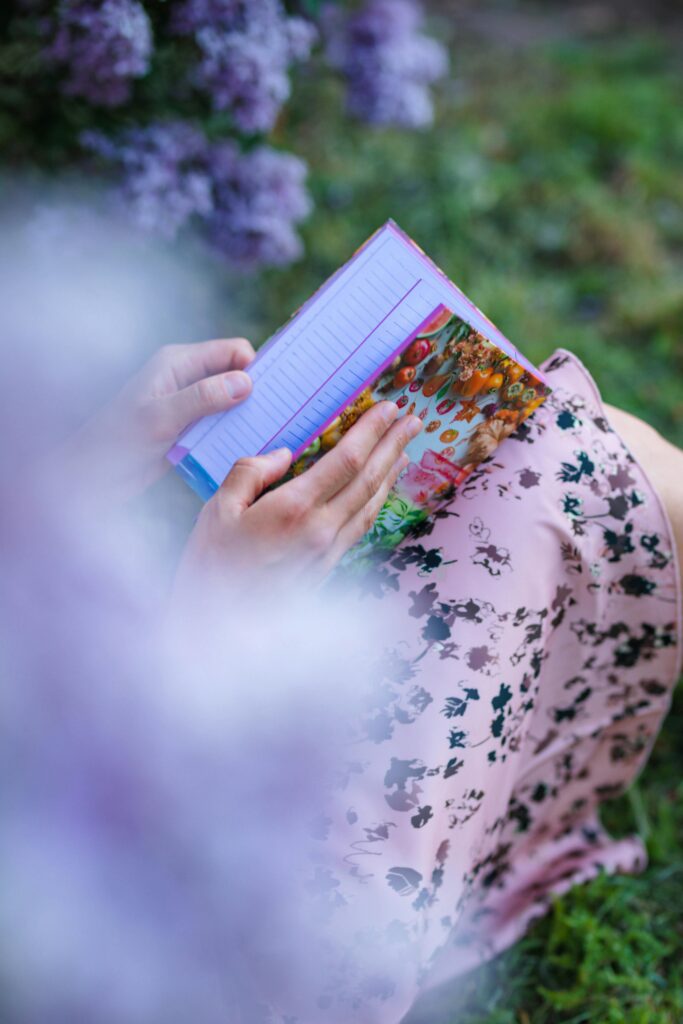 A serene moment captured as a woman reads a colorful notebook surrounded by lush greenery and purple flowers.